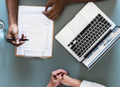 Two people in front of a computer with a clipboard and paperwork